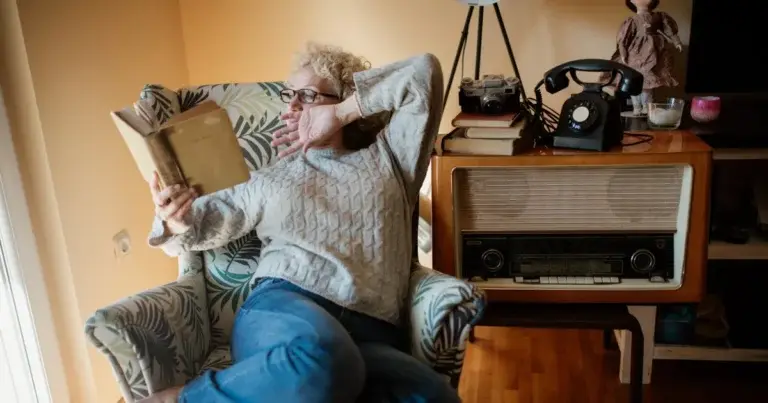 A woman stretches and yawns while reading in an armchair by a window
