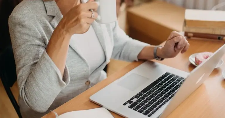 A woman sips coffee while working at a laptop with a notebook at a sunlit desk