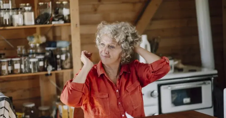 A woman lifts her curly hair off her neck while sitting in a bright kitchen