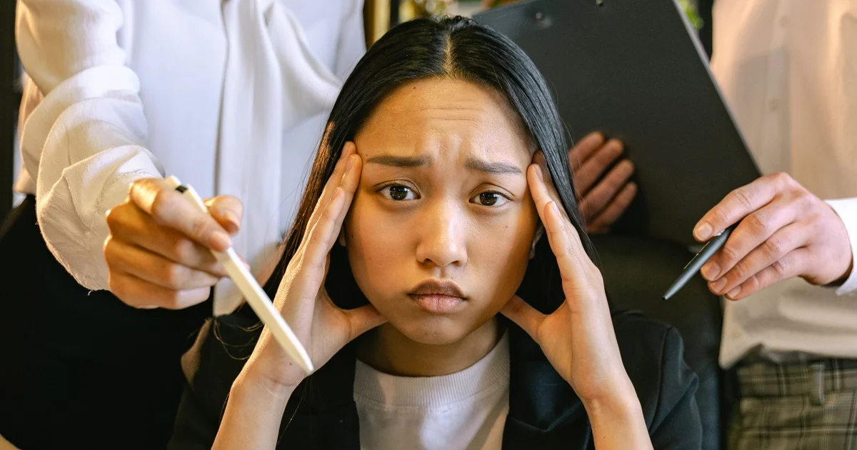Stressed woman at a desk pressing her hands to her temples while coworkers hold out papers and pens around her