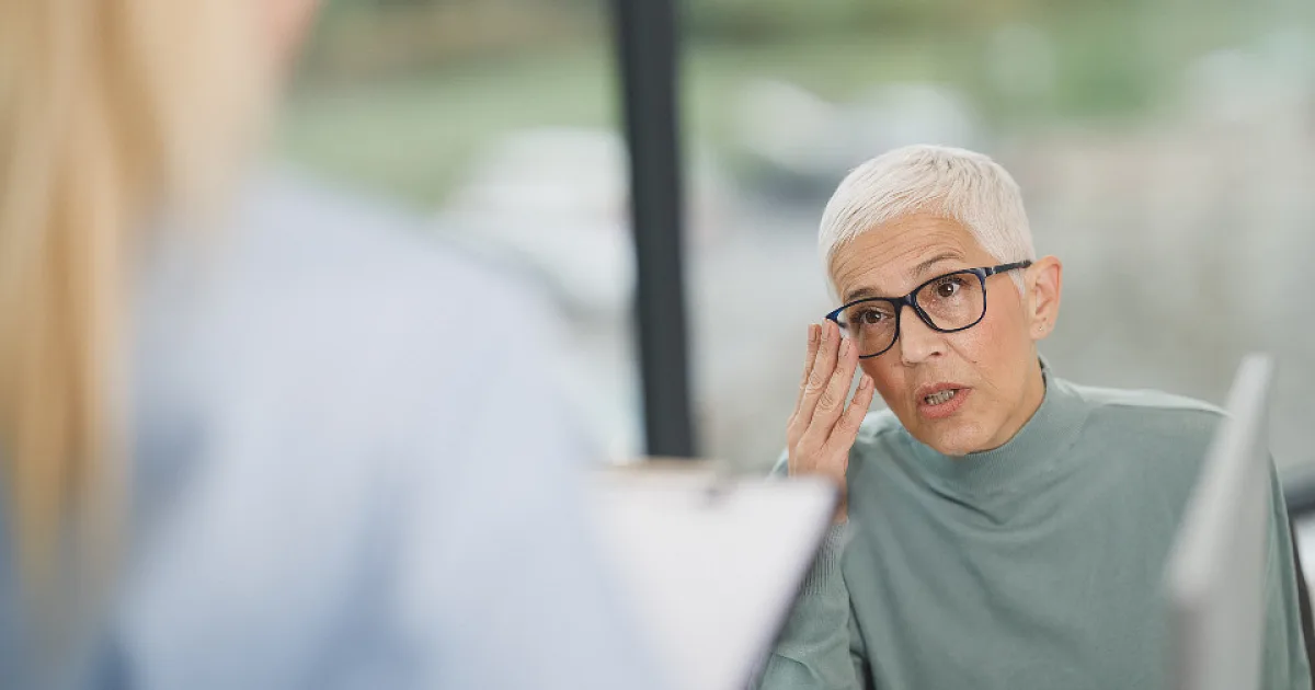 Woman with short grey hair and glasses describing her symptoms to a clinician during an appointment