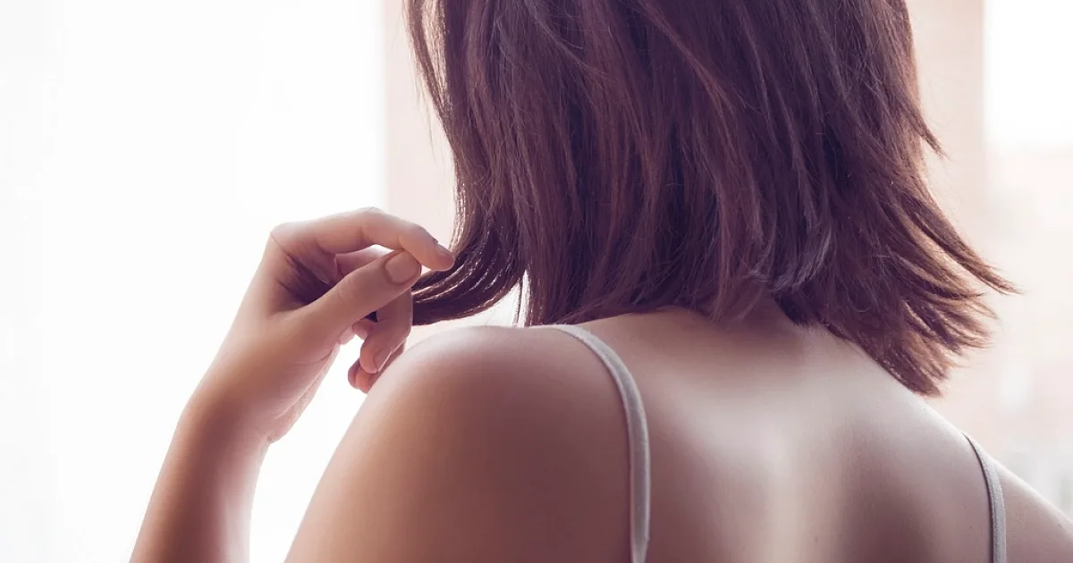 Woman seen from behind holding the ends of her shoulder-length hair by a bright window