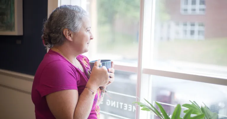 Woman in her fifties holding a mug of tea and looking calmly out of a window