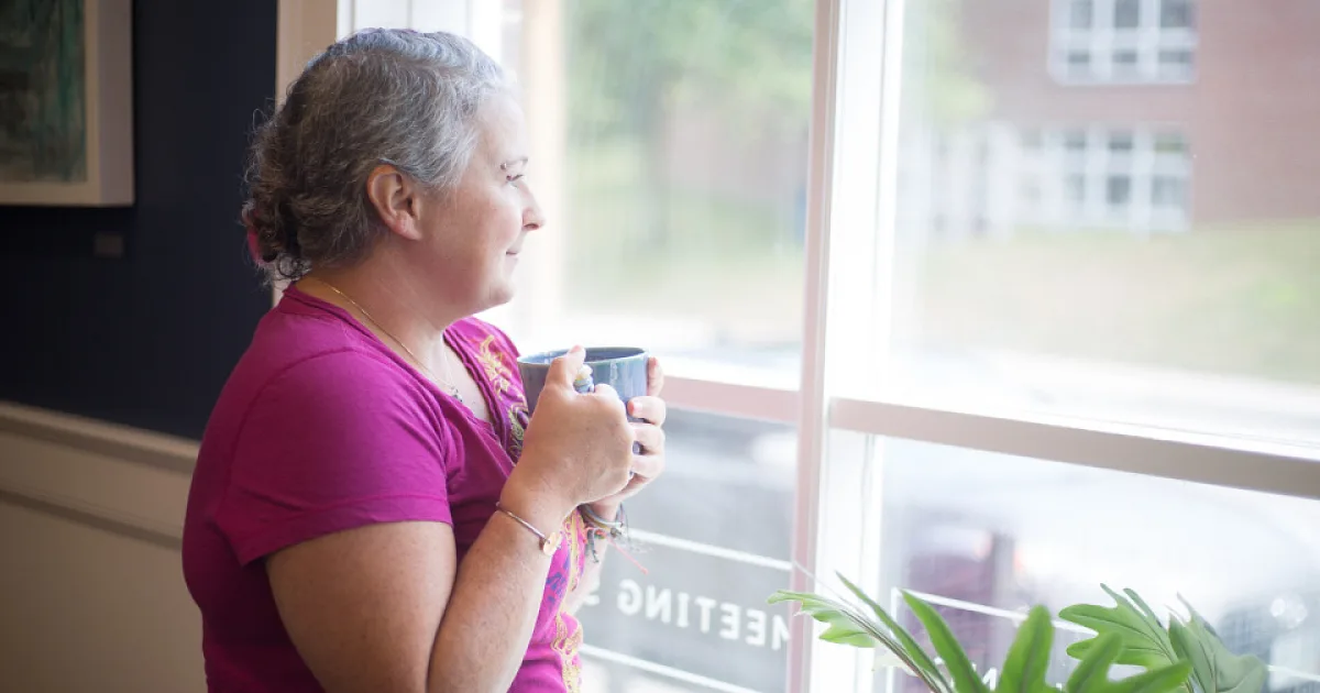 Woman in her fifties holding a mug of tea and looking calmly out of a window