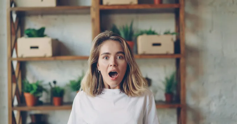 Surprised blonde woman with her mouth open, standing in front of shelves of potted plants