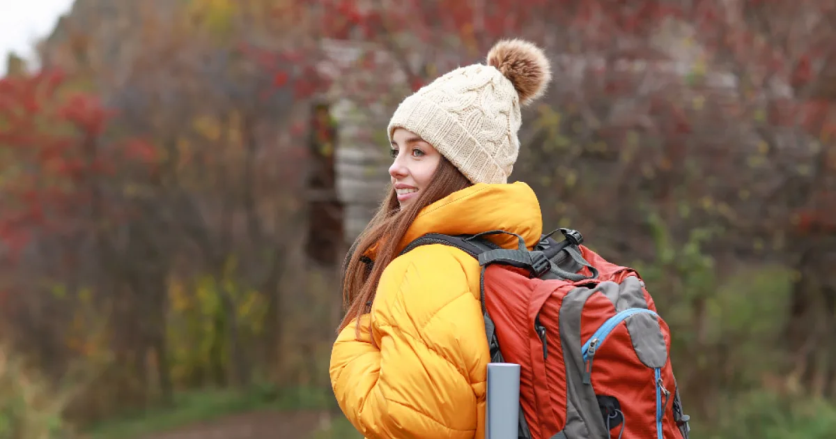 Smiling woman in a yellow jacket and knit hat with a backpack, out walking on an autumn trail