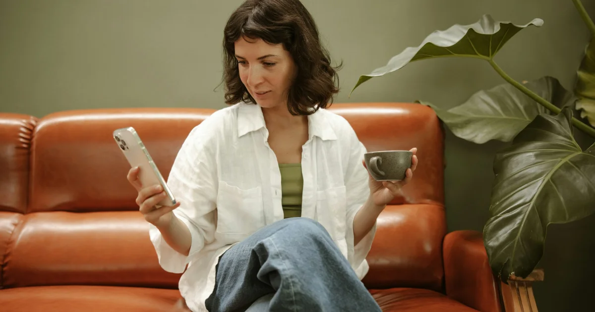 Woman in her forties sitting on a leather sofa with a cup of tea, looking at her phone
