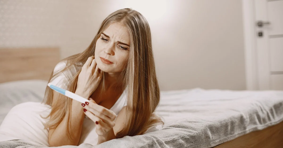 Woman lying on a bed looking worried at a pregnancy test in her hand